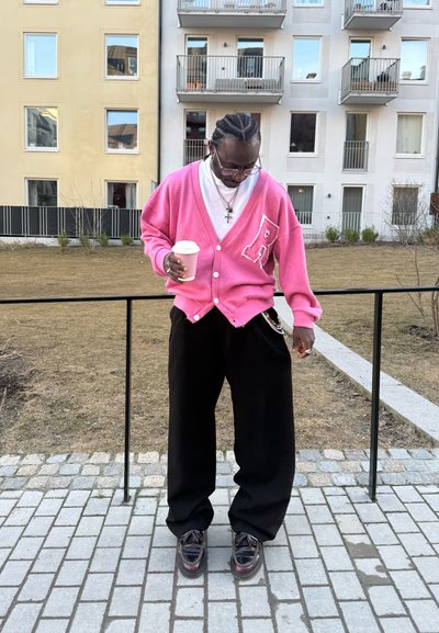 Hombre con cabello trenzado mira hacia abajo, lleva un cárdigan rosa, pantalones negros y gafas, sosteniendo una taza de café para llevar al aire libre frente a edificios.