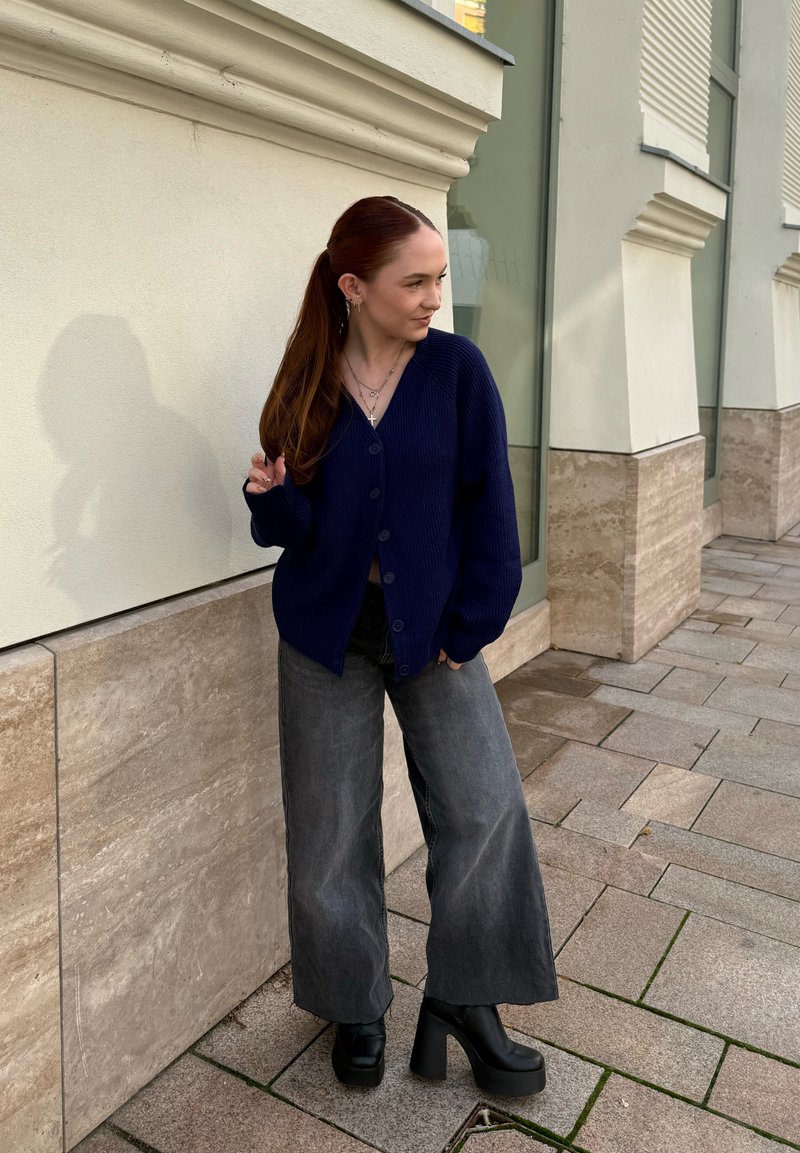 Cardigan en laine bleu marine avec fermeture à boutons, associé à un jean gris à jambes larges et des chaussures noires à plateforme épaisses. Surfaces texturées visibles.