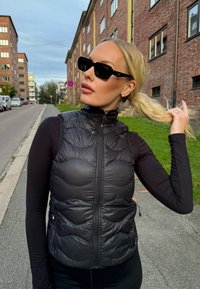 Μαύρο γιλέκο πούφ με ψηλό γιακά και με ανάγλυφη υφή, φορεμένο πάνω από μια μαύρη μπλούζα με μακριά μανίκια. Μαύρα τετράγωνα γυαλιά ηλίου ολοκληρώνουν την εμφάνιση.