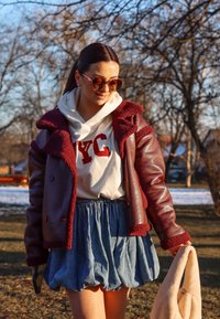 Burgundska usnjena jakna s sherpa podlogo, bela kapuca s rdečim tiskom "NYC", križan denim krilo, drži kremasto torbico.