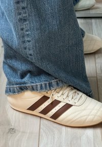 Sneakers bege claro com três riscas castanhas, couro texturizado e um design acolchoado, combinados com umas calças de ganga azuis.