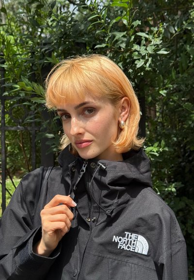 Joven mujer con pelo corto rubio ajustando el cuello de una chaqueta negra de The North Face, de pie al aire libre con vegetación de fondo.