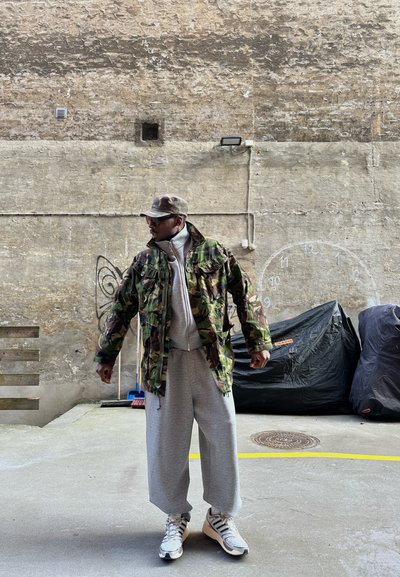 Hombre con chaqueta de camuflaje, pantalones deportivos grises y gorra, de pie junto a una pared de hormigón con un reloj dibujado con tiza y objetos cubiertos en un entorno urbano al aire libre.