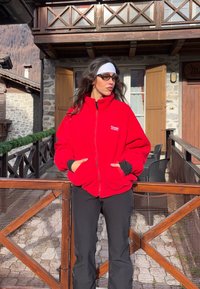 Femme portant une veste polaire rouge et un pantalon noir, debout sur un balcon en bois devant une maison en pierre avec des montagnes en arrière-plan.