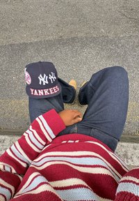 Navy Yankees cap with red lettering, red and blue striped jumper, and dark jeans. Smooth fabric texture, relaxed fit.
