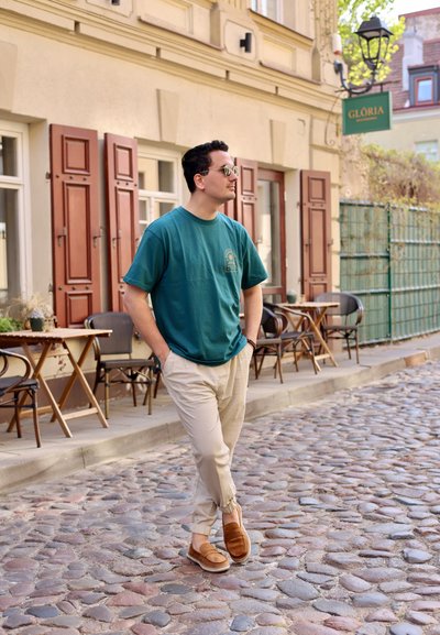 Hombre con gafas de sol, camisa turquesa y pantalones beiges caminando por una calle de adoquines frente a un café con mesas y sillas al aire libre.