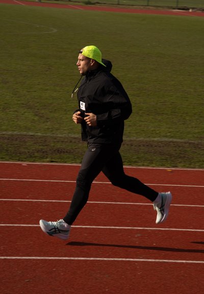 Corredor masculino con chaqueta negra y mallas ajustadas, gorra amarillo neón y zapatillas de correr azul claro, corriendo a toda velocidad en una pista roja.