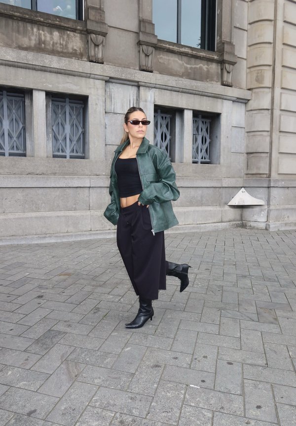 Woman wearing a green jacket, black crop top, wide black pants, boots, and sunglasses walking on a stone-paved street by a gray building.