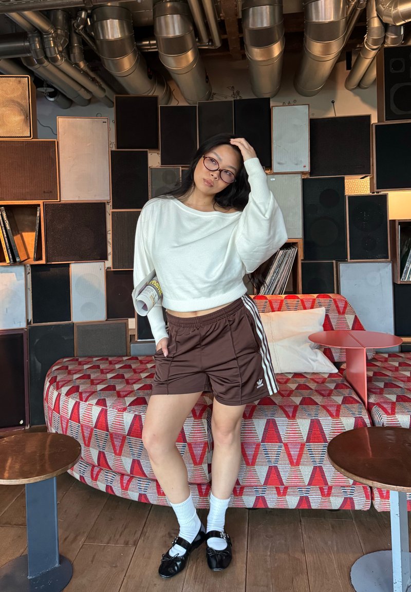 A woman wearing glasses, a white sweater, and brown shorts stands with one hand on her head in front of speakers and a patterned sofa.