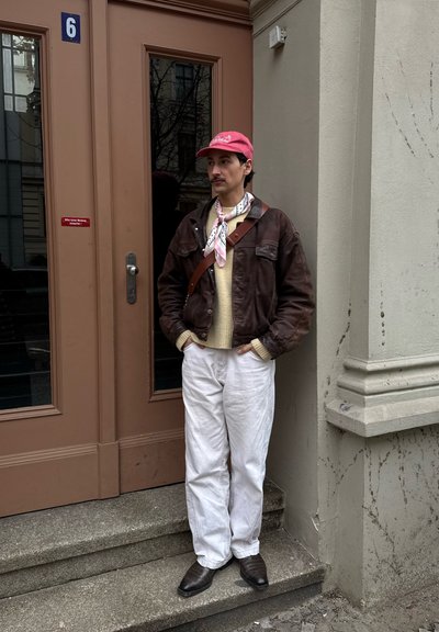 Hombre con gorra roja, chaqueta marrón, suéter crema, pantalones blancos y bufanda, de pie en las escaleras junto a una puerta marrón con el número 6 en el edificio.