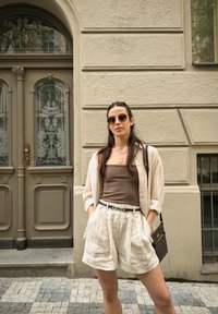 Young woman wearing sunglasses, striped shirt, brown tank top, and beige shorts stands with hands in pockets by an ornate wooden door on a city street.