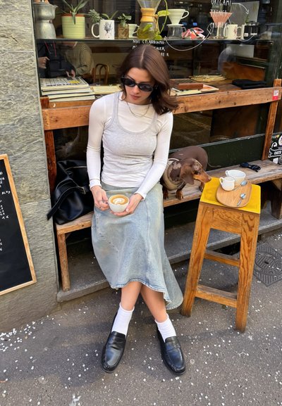 Mujer con gafas de sol sosteniendo un latte, sentada en un banco fuera de una cafetería con un perro pequeño y un taburete de madera que sostiene tazas de café y cucharas.