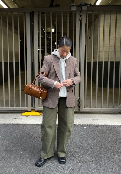 Blazer marrón de cuadros sobre sudadera con capucha gris, pantalones anchos color oliva y zapatos de plataforma negros. Bolso marrón con asas. Rejas al fondo.
