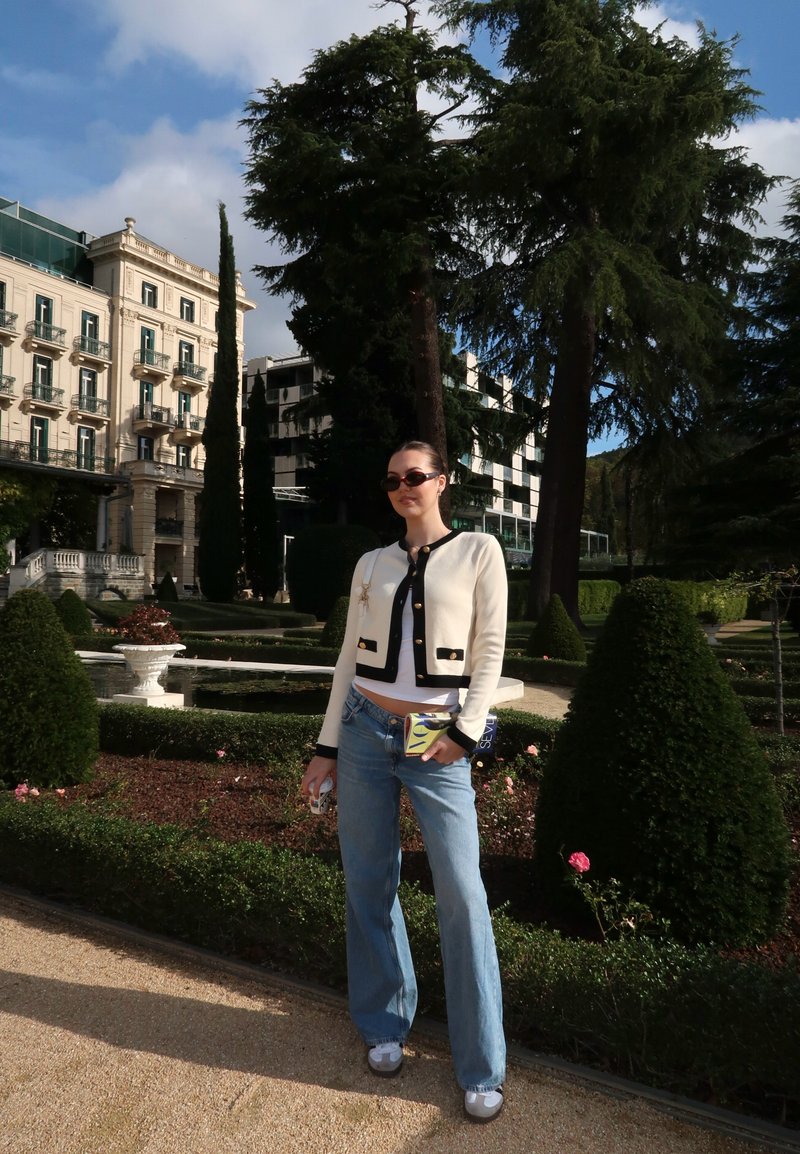 Cream cardigan with black details and gold buttons, paired with light blue wide-leg jeans. Model holds a colourful phone case, surrounded by greenery.