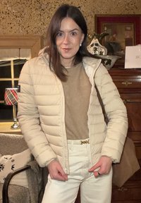 Doudoune beige clair avec coutures horizontales, portée sur un haut texturé couleur châtaigne, associée à un pantalon crème clair et une fermeture de ceinture dorée.