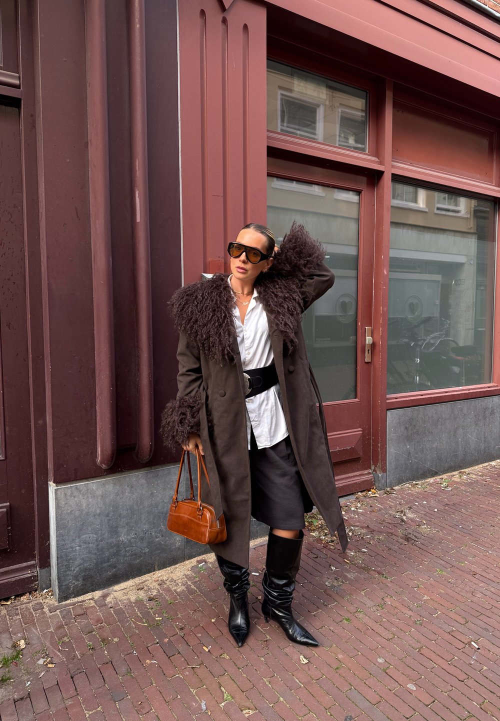 Femme portant un manteau sombre bordé de fourrure, une chemise blanche, une jupe noire, des bottes et des lunettes de soleil, debout sur un trottoir en briques, tenant un sac à main marron.