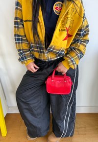 Gul rutete glidelåsjakke med rødt logo, svarte nylonbukser med hvite striper, holder en liten rød håndveske med logo. Tre gulvbakgrunn.