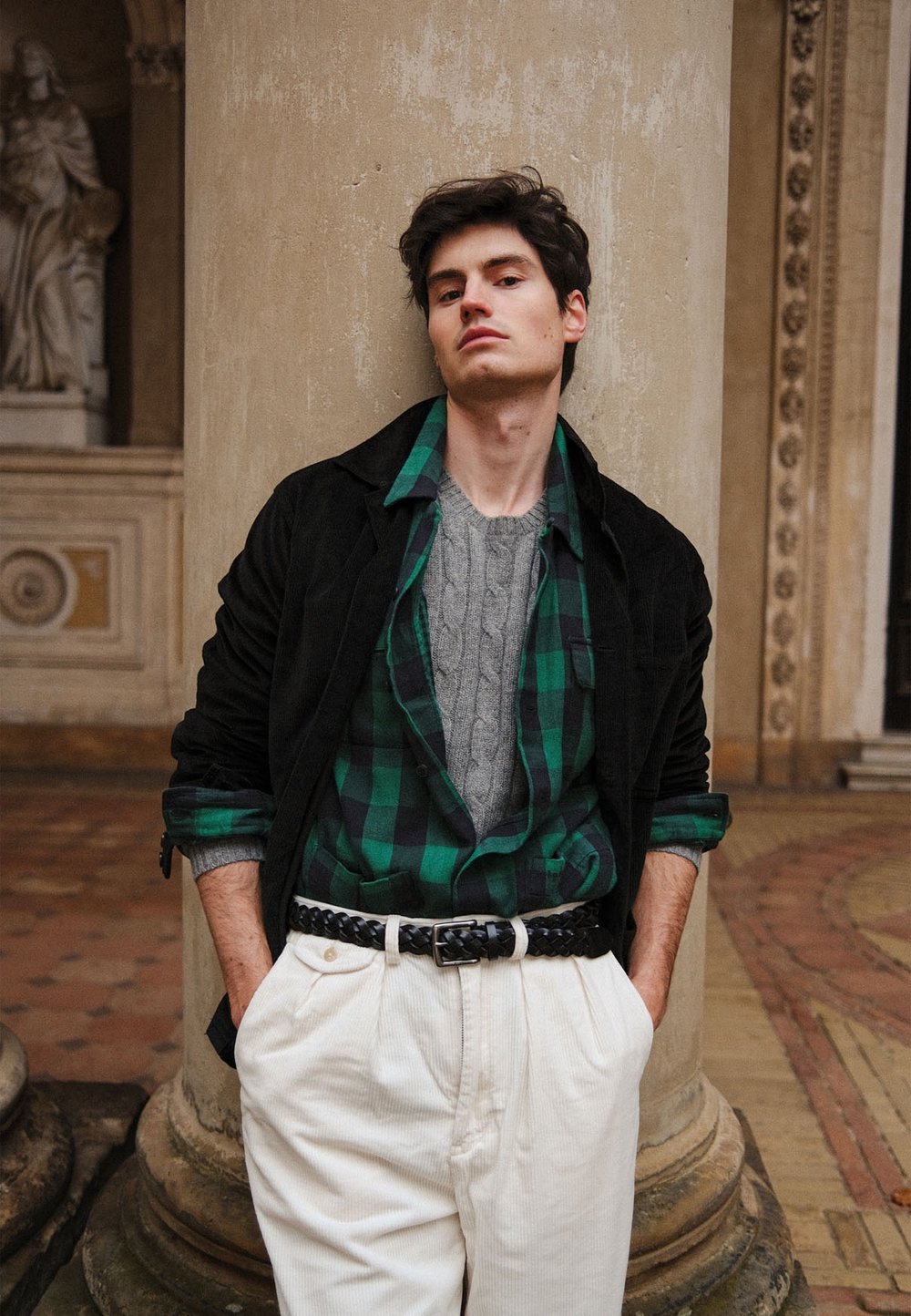 Veste en velours côtelé noire, chemise en flanelle à carreaux verts, pull gris à torsades, pantalon en velours côtelé blanc, ceinture noire tissée, le tout posé contre un pilier en pierre.