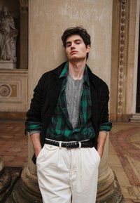 Veste en velours côtelé noire, chemise en flanelle à carreaux verts, pull gris à torsades, pantalon en velours côtelé blanc, ceinture noire tissée, le tout posé contre un pilier en pierre.