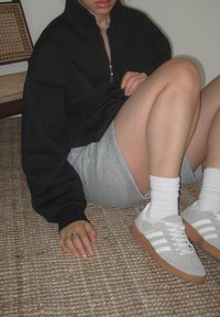 Άτομο που κάθεται στο χαλί, φορόντας μαύρη φούτερ με φερμουάρ, γκρίζα σορτς, λευκές κάλτσες και γκρίζα αθλητικά παπούτσια Adidas με καφέ σόλες.