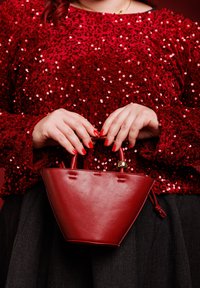 Bolso de cuero rojo con una forma estructurada y triangular, que cuenta con un asa corta y un detalle dorado. Combinado con una blusa roja con lentejuelas y una falda oscura.