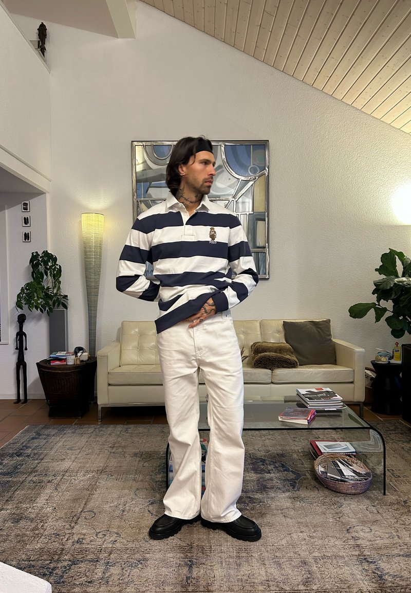 Polo à manches longues rayé bleu marine et blanc, associé à un pantalon large blanc et des chaussures noires. Cadre intérieur avec une décoration minimaliste.