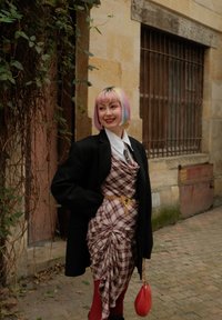 Ung person med pastelfarvet pink og blå hår iført ternet kjole, sort jakke, guld kædebælte, røde strømpebukser og holder en rød taske nær en gammel stenbygning.
