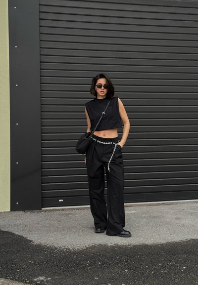 Top corto negro, pantalones anchos negros con detalle de cadena, bolso negro, gafas de sol negras, de pie frente a una puerta de garaje enrollable gris.