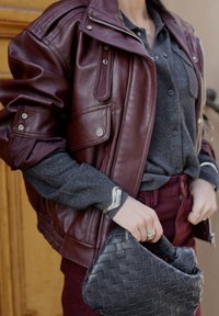 Burgundy leather bomber jacket with a shiny finish, paired with a gray sweater and dark red pants, holding a black woven handbag.