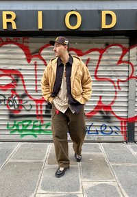 Χακί μπουφάν με κουκούλα πάνω από ένα σκούρο πουκάμισο, συνδυασμένο με ριγέ πουκάμισο και καφέ παντελόνι. Μαύρα παπούτσια με υφή; φόντο με γκράφιτι.