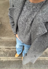 Manteau en laine grise avec une finition texturée, superposé à un pull en maille foncé, assorti à un jean ample bleu clair et des chaussures marron clair.