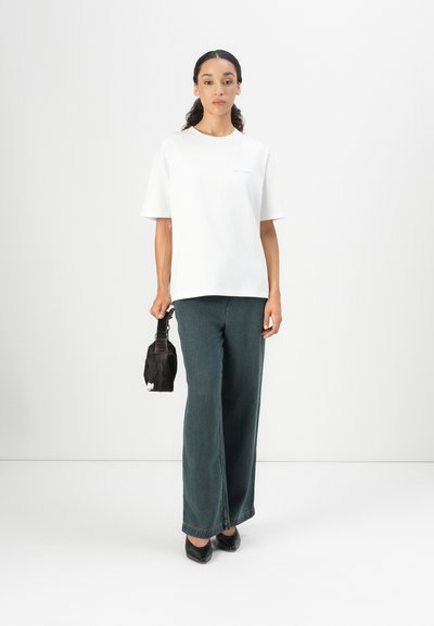 Camiseta blanca de algodón con mangas cortas, combinada con unos pantalones anchos de mezclilla verde oscuro. Modelo sosteniendo un pequeño bolso negro, usando tacones negros.