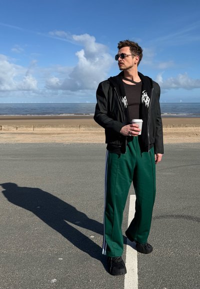 Hombre con gafas de sol que lleva una chaqueta negra y pantalones deportivos verdes sostiene una taza de café, de pie en una carretera junto a la playa bajo un cielo azul.
