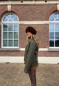 Groene leren jas met een losse pasvorm, een bruine bonten hoed en een bruine corduroy rok, gecombineerd met bruine kniehoge laarzen. Bakstenen muur op de achtergrond.
