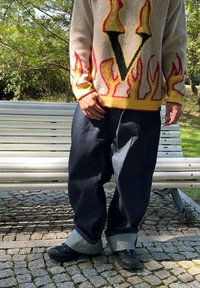 Μπεζ πλεκτό πουλόβερ με σχέδια φλόγας και κεντρικό μαύρο "V", συνδυασμένο με φαρδιά μπλε τζιν παντελόνια με γυρισμένα τελειώματα και μαύρα υποδήματα.