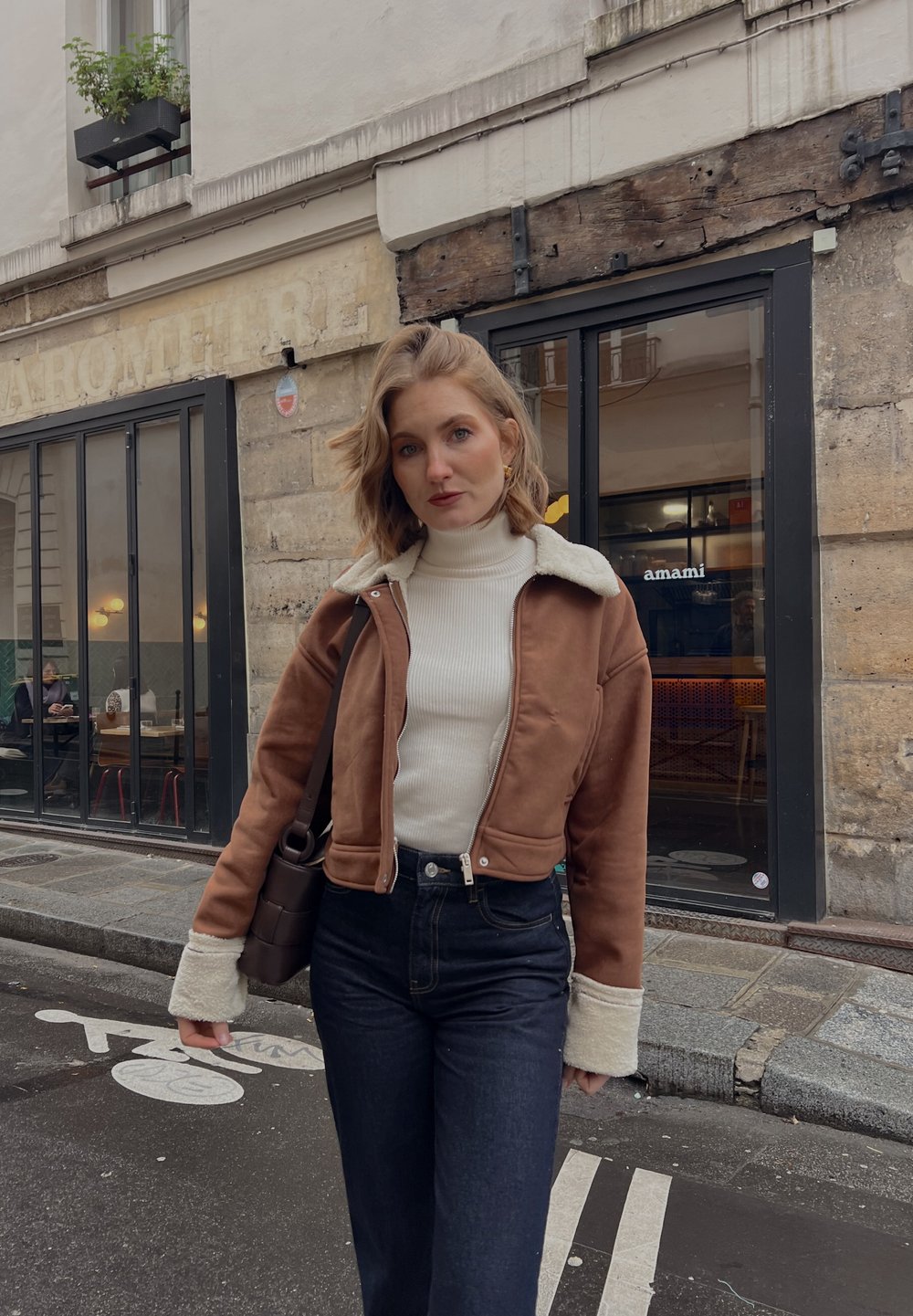 Bruine suède cropped jas met een witte schapenvacht kraag, gecombineerd met een crèmekleurige coltrui en hooggewaisted donkerdenim jeans. Bruine schoudertas.