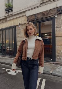 Bruine suède cropped jas met een witte schapenvacht kraag, gecombineerd met een crèmekleurige coltrui en hooggewaisted donkerdenim jeans. Bruine schoudertas.