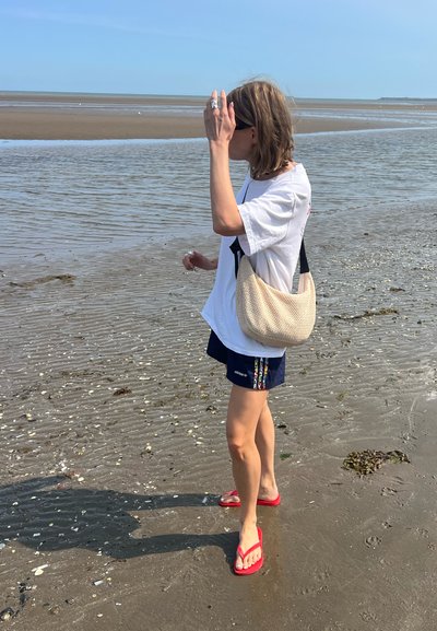 Persona de pie en arena mojada, vistiendo una camiseta blanca, pantalones cortos marinos con rayas coloridas y chanclas rojas, sosteniendo una bolsa beige.