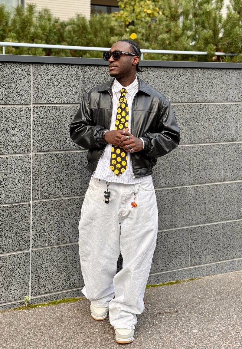 Veste en cuir noir, chemise blanche à rayures, cravate jaune avec un visage souriant, pantalon blanc ample et baskets ; accessoires comprenant des breloques et des bagues.