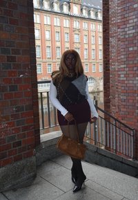 Brązowa i szara dzianinowa kamizelka na biały, długi rękaw, do tego bordowa mini spódnica i czarne rajstopy. Trzyma brązową torebkę, ma na sobie szpilki.