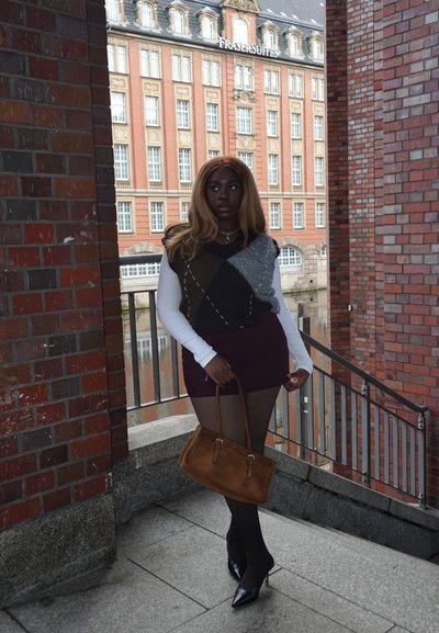 Chaleco de punto marrón y gris sobre una camisa blanca de manga larga, combinado con una minifalda burdeos y medias negras. Sosteniendo un bolso marrón, con tacones.