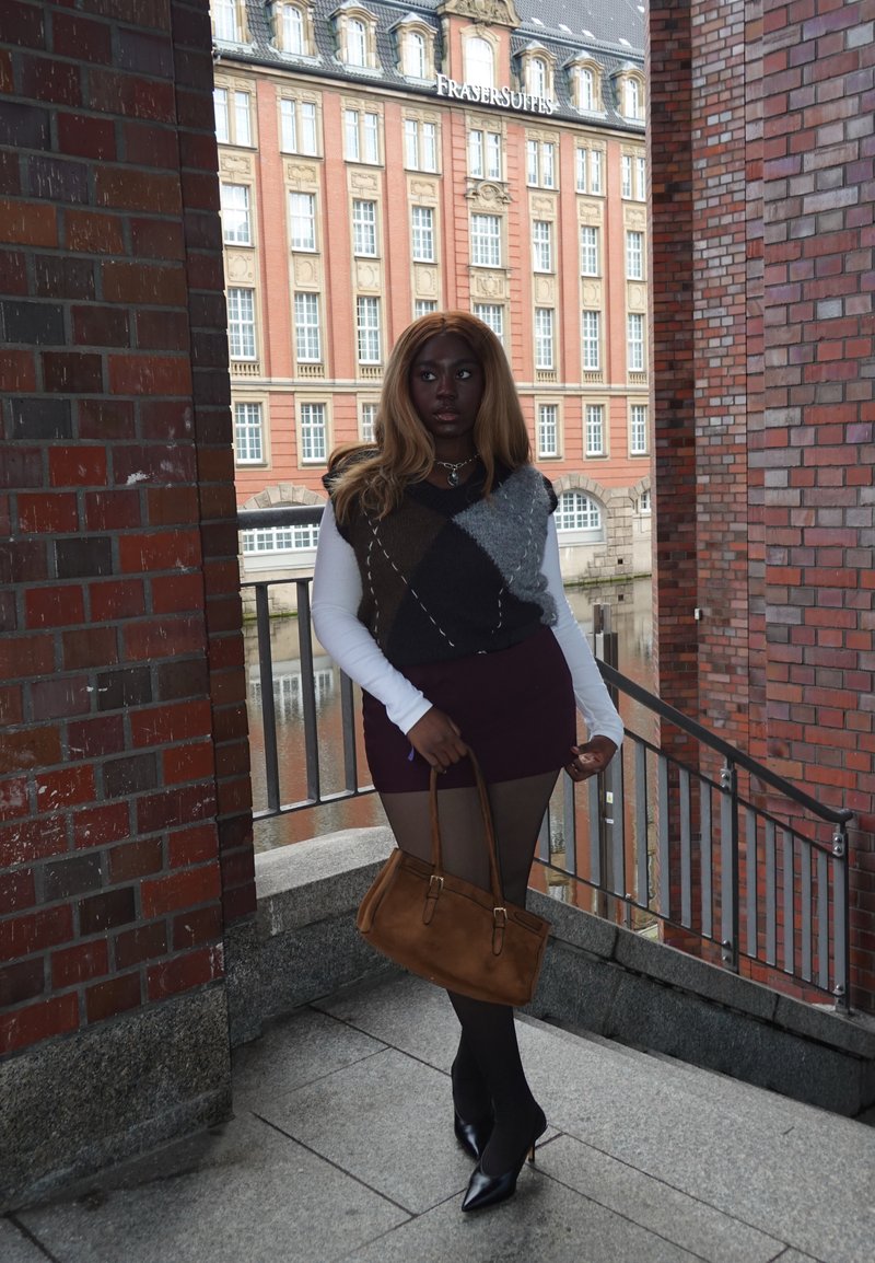 Veste en tricot marron et grise portée sur une chemise blanche à manches longues, associée à une mini-jupe bordeaux et des collants noirs. Tenant un sac à main marron, portant des talons.