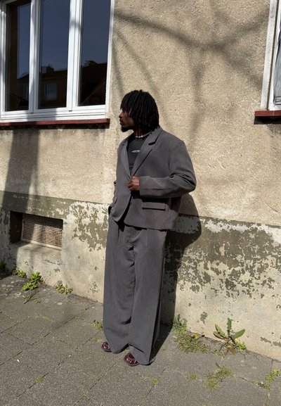 Hombre con rastas lleva un traje gris sobredimensionado y camisa negra, de pie contra una pared beige desgastada con ventanas en un día soleado.