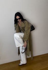 Gabardina caqui, camiseta gris, jeans de pierna ancha blancos, gafas de sol negras y zapatillas grises con rayas negras. Fondo de suelo de madera.