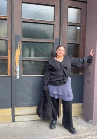 Fekete csíkos blúz fodros ujjal, réteges kék mintás szoknya, fekete bő szabású nadrág és fekete platform cipő. Szőr kiegészítővel rendelkezik.