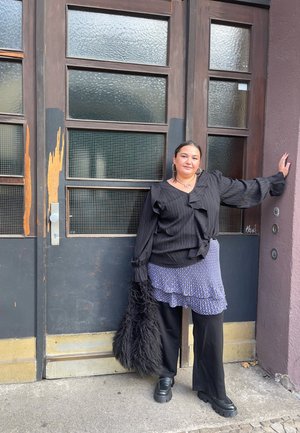 Blusa a rayas negras con mangas acampanadas, falda de capas azul con patrón, pantalones anchos negros y zapatos de plataforma negros. Presenta un acento de piel.