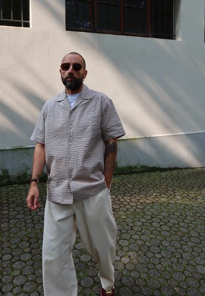 Camisa beige de manga corta con patrón texturado, combinada con pantalones sueltos de color marfil y zapatos burdeos, sobre un fondo de pared de color claro.