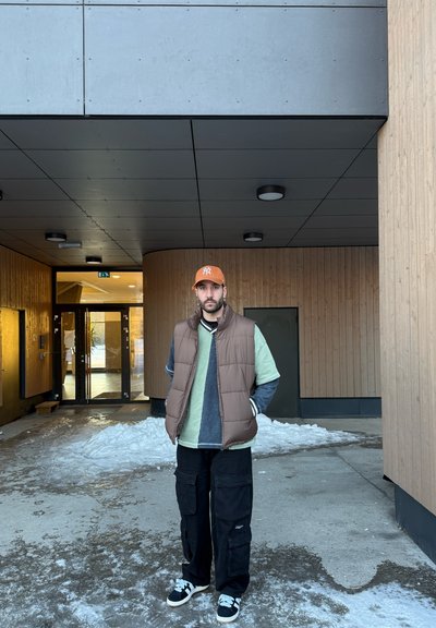 Chaleco de plumas marrón sobre una camisa verde, pantalones cargo negros y zapatillas negras. Gorra naranja con logo. Entorno al aire libre con elementos de concreto y madera.