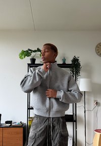 Sweat-shirt gris à fermeture éclair avec un col montant et des poignets épais, porté par-dessus un pantalon cargo décontracté de couleur foncée, sur un fond intérieur minimaliste.