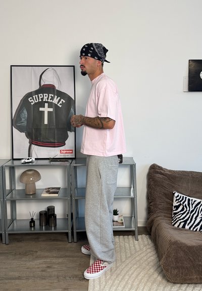 Hombre con bandana, camisa rosa y pantalones grises posa junto a estanterías metálicas sosteniendo un póster enmarcado de una chaqueta Supreme con una cruz en la espalda.